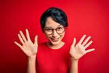 Young beautiful asian girl wearing casual t-shirt and glasses over isolated red background showing and pointing up with fingers number ten while smiling confident and happy.