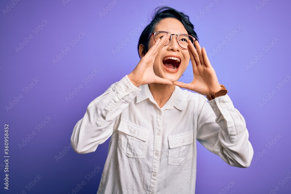 Young beautiful asian girl wearing casual shirt and glasses standing over purple background Shouting angry out loud with hands over mouth Young beautiful asian girl wearing casual shirt and glasses standing over purple background Shouting angry out loud with hands over mouth