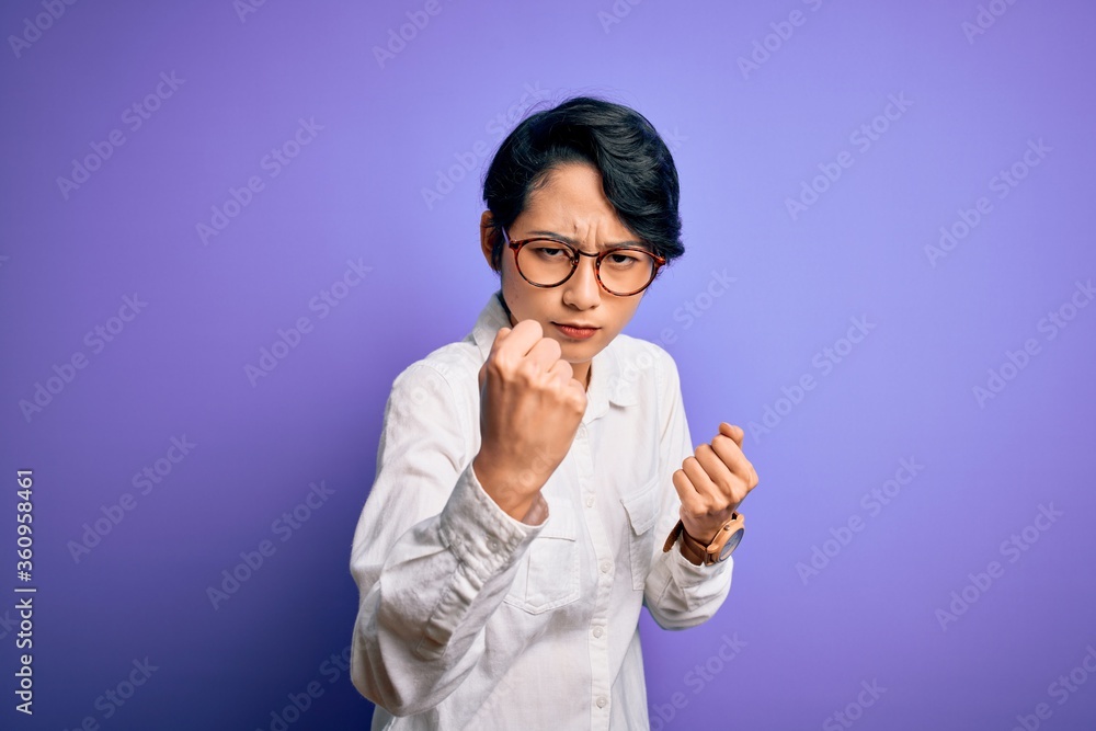 Young beautiful asian girl wearing casual shirt and glasses standing over purple background Ready to fight with fist defense gesture, angry and upset face, afraid of problem Young beautiful asian girl wearing casual shirt and glasses standing over purple background Ready to fight with fist defense gesture, angry and upset face, afraid of problem