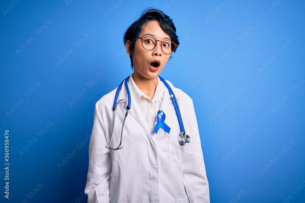 Young beautiful asian doctor girl wearing stethoscope and coat with blue cancer ribbon afraid and shocked with surprise expression, fear and excited face. Young beautiful asian doctor girl wearing stethoscope and coat with blue cancer ribbon afraid and shocked with surprise expression, fear and excited face.