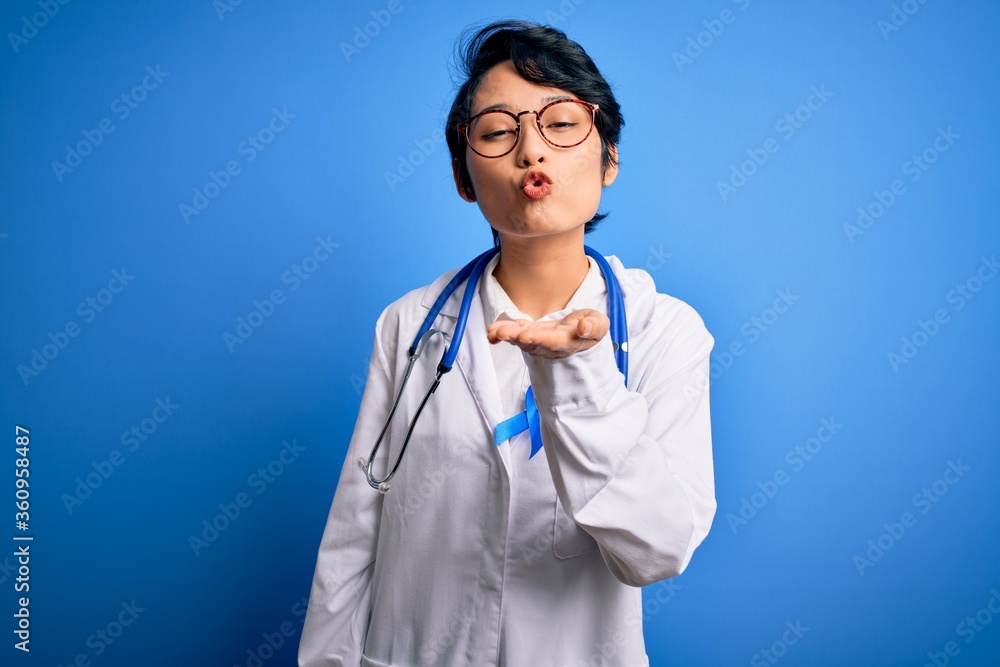 Young beautiful asian doctor girl wearing stethoscope and coat with blue cancer ribbon looking at the camera blowing a kiss with hand on air being lovely and sexy. Love expression. Young beautiful asian doctor girl wearing stethoscope and coat with blue cancer ribbon looking at the camera blowing a kiss with hand on air being lovely and sexy. Love expression.