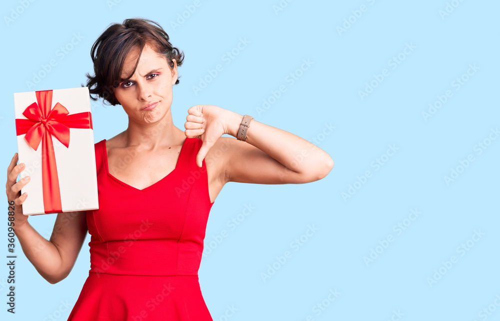 Beautiful young woman with short hair holding gift with angry face, negative sign showing dislike with thumbs down, rejection concept Beautiful young woman with short hair holding gift with angry face, negative sign showing dislike with thumbs down, rejection concept