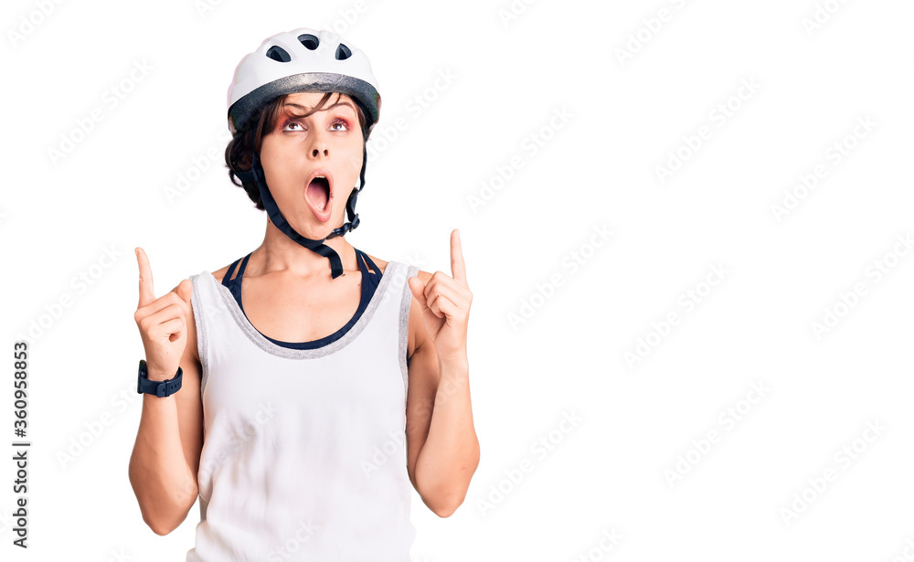 Beautiful young woman with short hair wearing bike helmet amazed and surprised looking up and pointing with fingers and raised arms. Beautiful young woman with short hair wearing bike helmet amazed and surprised looking up and pointing with fingers and raised arms.