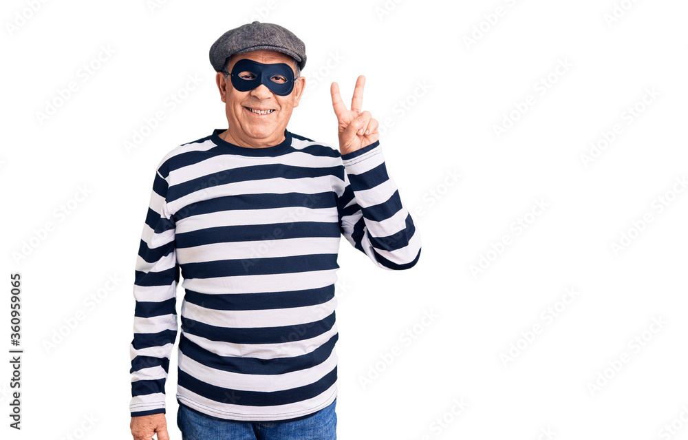 Fototapeta premium Senior handsome man wearing burglar mask and t-shirt smiling with happy face winking at the camera doing victory sign with fingers. number two.