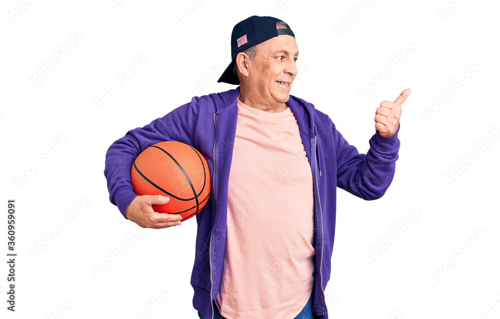 Senior handsome grey-haired man holding basketball ball pointing thumb up to the side smiling happy with open mouth Senior handsome grey-haired man holding basketball ball pointing thumb up to the side smiling happy with open mouth