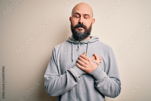 Handsome sporty bald man with beard wearing sweatshirt standing over pink background smiling with hands on chest with closed eyes and grateful gesture on face. Health concept.