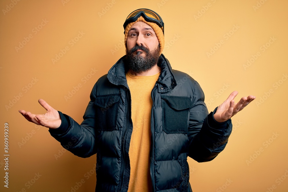 Handsome skier bald man with beard skiing wearing snow sportswear and ski goggles clueless and confused expression with arms and hands raised. Doubt concept. Handsome skier bald man with beard skiing wearing snow sportswear and ski goggles clueless and confused expression with arms and hands raised. Doubt concept.
