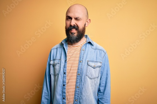 Handsome bald man with beard wearing casual denim jacket and striped t-shirt winking looking at the camera with sexy expression, cheerful and happy face.