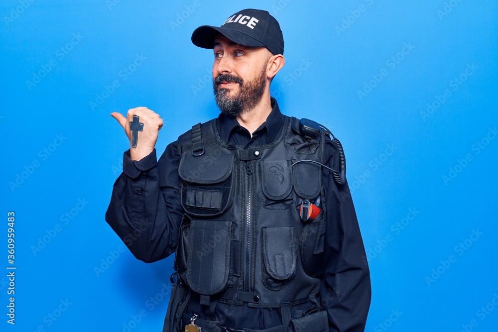 Young handsome man wearing police uniform smiling with happy face looking and pointing to the side with thumb up. Young handsome man wearing police uniform smiling with happy face looking and pointing to the side with thumb up.
