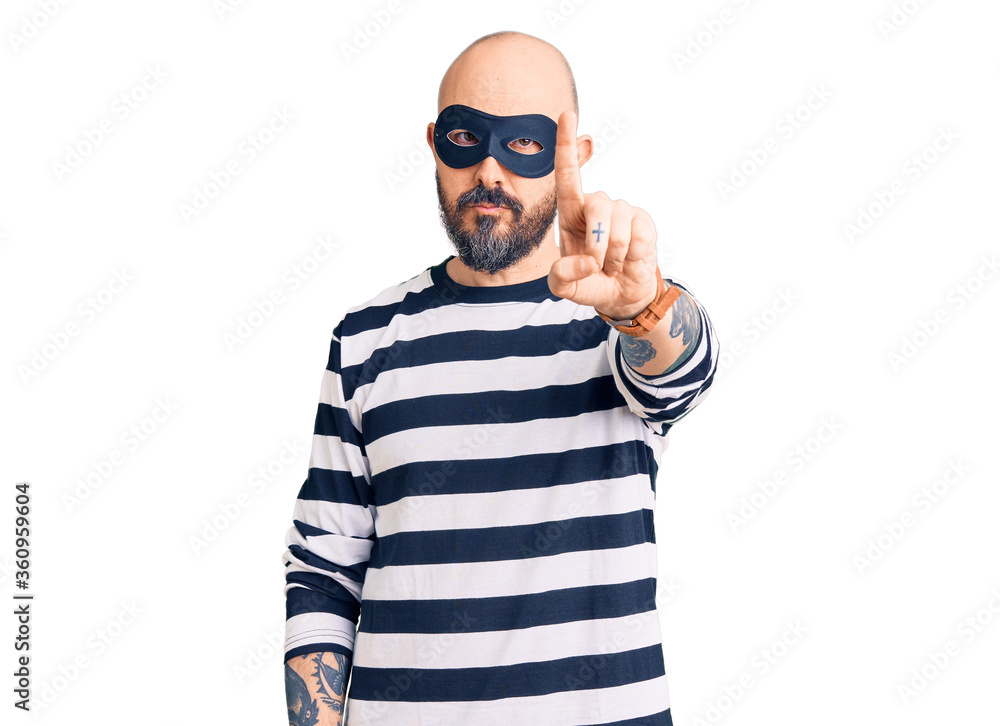 Young handsome man wearing burglar mask pointing with finger up and angry expression, showing no gesture Young handsome man wearing burglar mask pointing with finger up and angry expression, showing no gesture