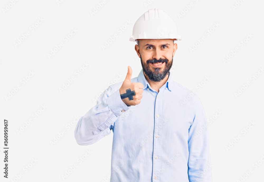 Young handsome man wearing architect hardhat doing happy thumbs up gesture with hand. approving expression looking at the camera showing success. Young handsome man wearing architect hardhat doing happy thumbs up gesture with hand. approving expression looking at the camera showing success.