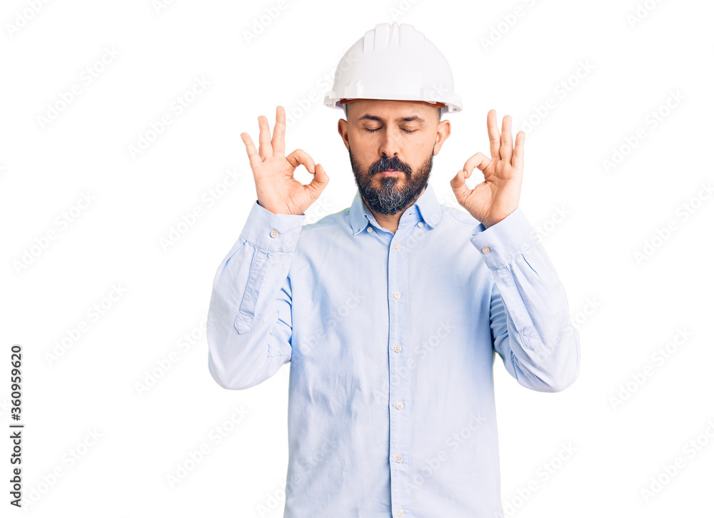 Young handsome man wearing architect hardhat relax and smiling with eyes closed doing meditation gesture with fingers. yoga concept. Young handsome man wearing architect hardhat relax and smiling with eyes closed doing meditation gesture with fingers. yoga concept.