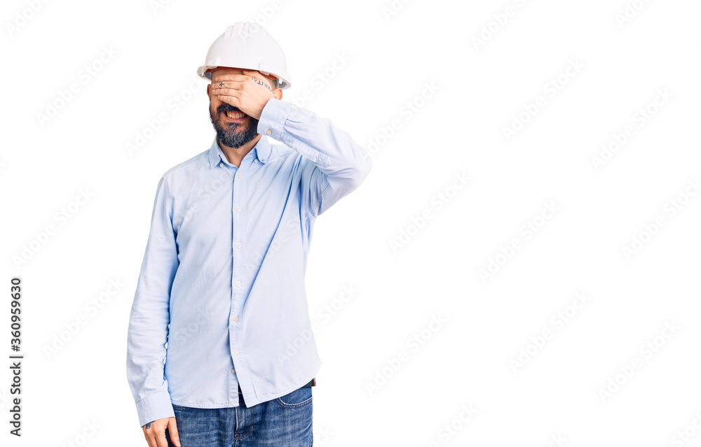 Young handsome man wearing architect hardhat smiling and laughing with hand on face covering eyes for surprise. blind concept. Young handsome man wearing architect hardhat smiling and laughing with hand on face covering eyes for surprise. blind concept.