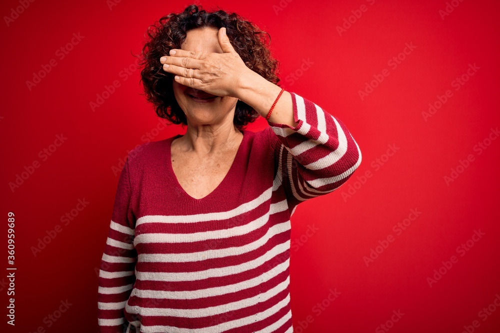 Middle age beautiful curly hair woman wearing casual striped sweater over red background smiling and laughing with hand on face covering eyes for surprise. Blind concept. Middle age beautiful curly hair woman wearing casual striped sweater over red background smiling and laughing with hand on face covering eyes for surprise. Blind concept.