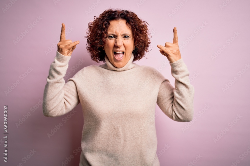 Middle age beautiful curly hair woman wearing casual turtleneck sweater over pink background shouting with crazy expression doing rock symbol with hands up. Music star. Heavy music concept. Middle age beautiful curly hair woman wearing casual turtleneck sweater over pink background shouting with crazy expression doing rock symbol with hands up. Music star. Heavy music concept.