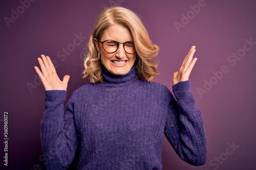 Middle age beautiful blonde woman wearing casual purple turtleneck sweater and glasses celebrating mad and crazy for success with arms raised and closed eyes screaming excited. Winner concept