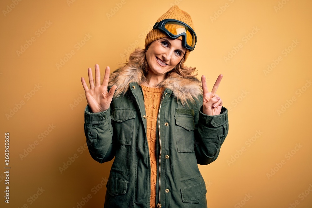 Middle age beautiful blonde skier woman wearing snow sportwear and ski goggles showing and pointing up with fingers number seven while smiling confident and happy.