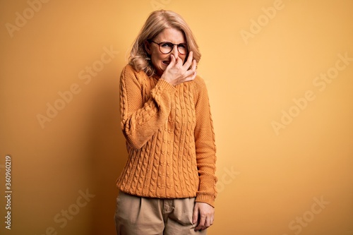 Middle age beautiful blonde woman wearing casual sweater and glasses over yellow background smelling something stinky and disgusting, intolerable smell, holding breath with fingers on nose. Bad smell © Krakenimages.com
