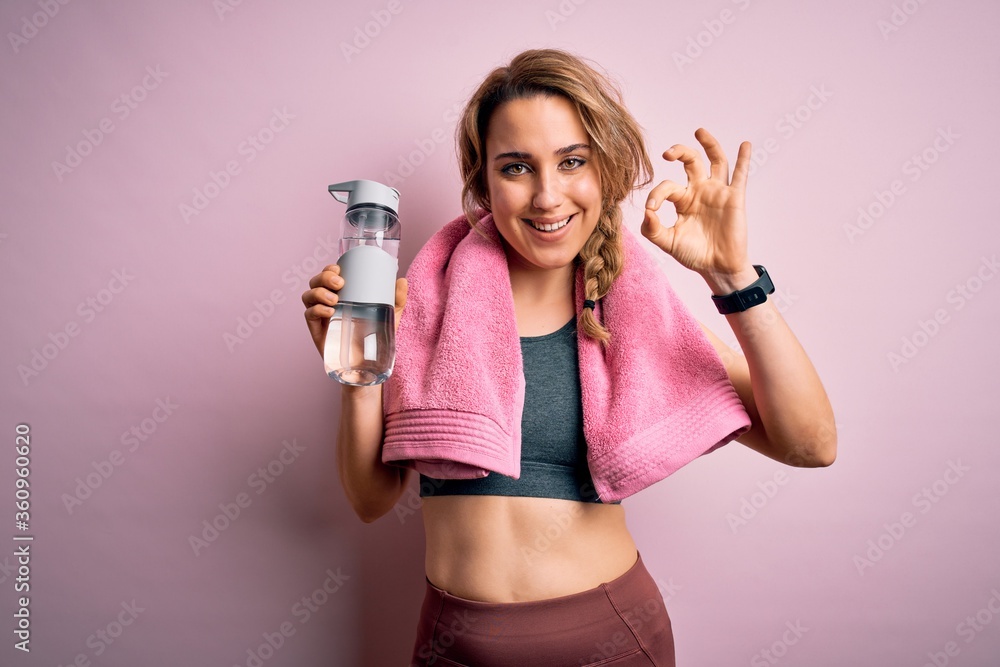Young beautiful blonde sportswoman wearing sportswear and towel drinking bottle of water doing ok sign with fingers, excellent symbol Young beautiful blonde sportswoman wearing sportswear and towel drinking bottle of water doing ok sign with fingers, excellent symbol