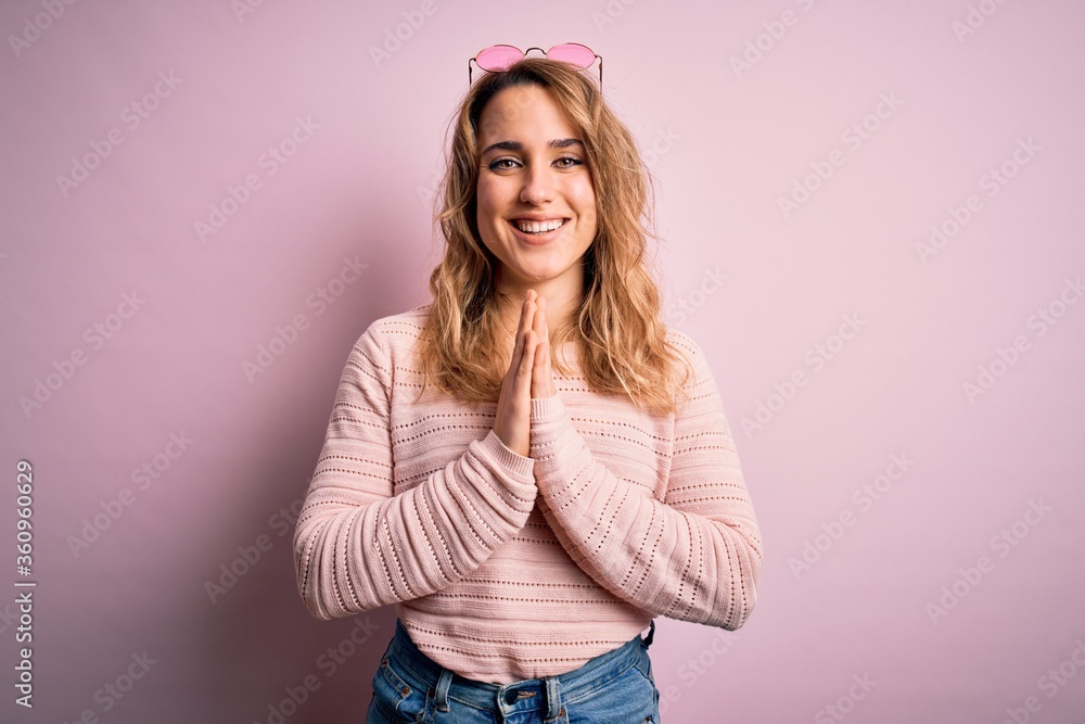 Fototapeta premium Young beautiful blonde woman wearing casual sweater and sunglasses over pink background praying with hands together asking for forgiveness smiling confident.