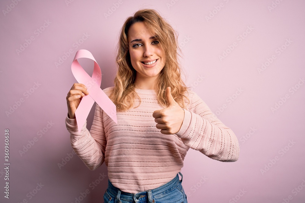 Young beautiful blonde woman holding pink cancer ribbon symbol over isolated background happy with big smile doing ok sign, thumb up with fingers, excellent sign Young beautiful blonde woman holding pink cancer ribbon symbol over isolated background happy with big smile doing ok sign, thumb up with fingers, excellent sign
