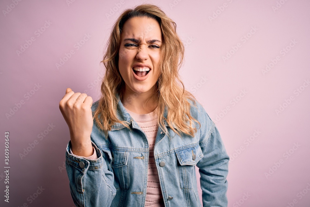 Young beautiful blonde woman wearing casual denim jacket standing over pink background angry and mad raising fist frustrated and furious while shouting with anger. Rage and aggressive concept. Young beautiful blonde woman wearing casual denim jacket standing over pink background angry and mad raising fist frustrated and furious while shouting with anger. Rage and aggressive concept.