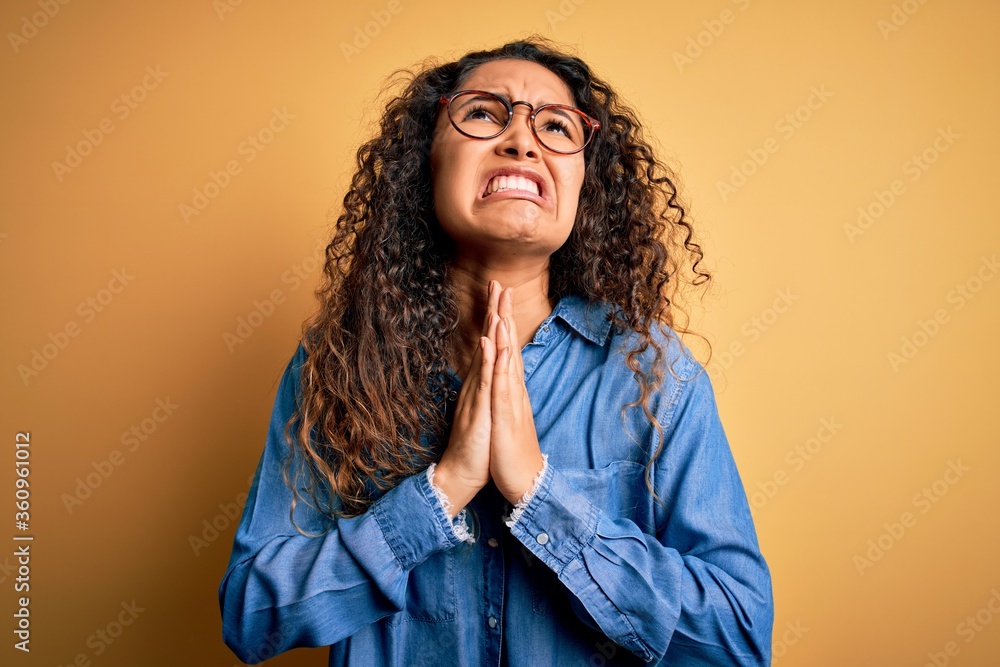 Beautiful woman with curly hair wearing casual denim shirt and glasses over yellow background begging and praying with hands together with hope expression on face very emotional and worried. Begging. Beautiful woman with curly hair wearing casual denim shirt and glasses over yellow background begging and praying with hands together with hope expression on face very emotional and worried. Begging.