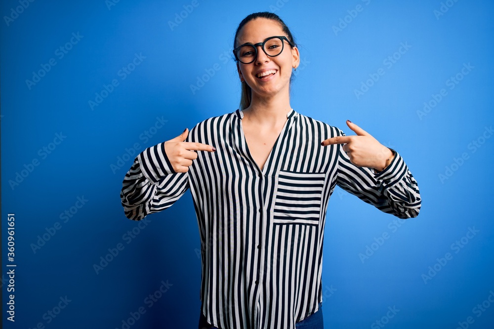 Beautiful blonde woman with blue eyes wearing striped shirt and glasses over blue background looking confident with smile on face, pointing oneself with fingers proud and happy. Beautiful blonde woman with blue eyes wearing striped shirt and glasses over blue background looking confident with smile on face, pointing oneself with fingers proud and happy.