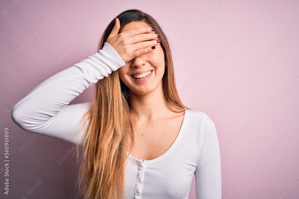 Young beautiful blonde woman with blue eyes wearing white t-shirt over pink background smiling and laughing with hand on face covering eyes for surprise. Blind concept. Young beautiful blonde woman with blue eyes wearing white t-shirt over pink background smiling and laughing with hand on face covering eyes for surprise. Blind concept.