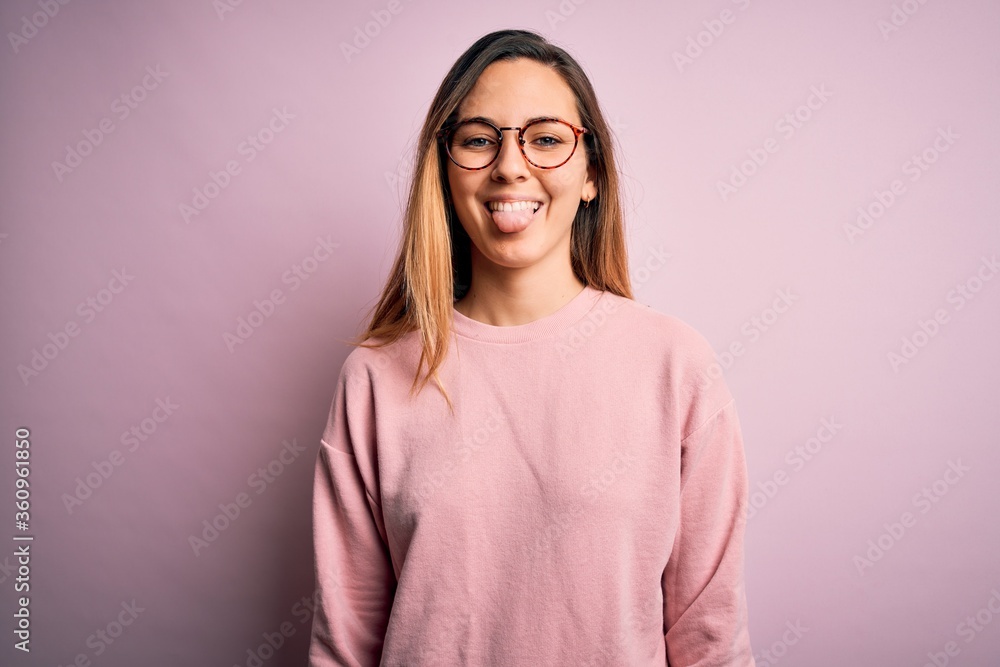 Beautiful blonde woman with blue eyes wearing sweater and glasses over pink background sticking tongue out happy with funny expression. Emotion concept. Beautiful blonde woman with blue eyes wearing sweater and glasses over pink background sticking tongue out happy with funny expression. Emotion concept.