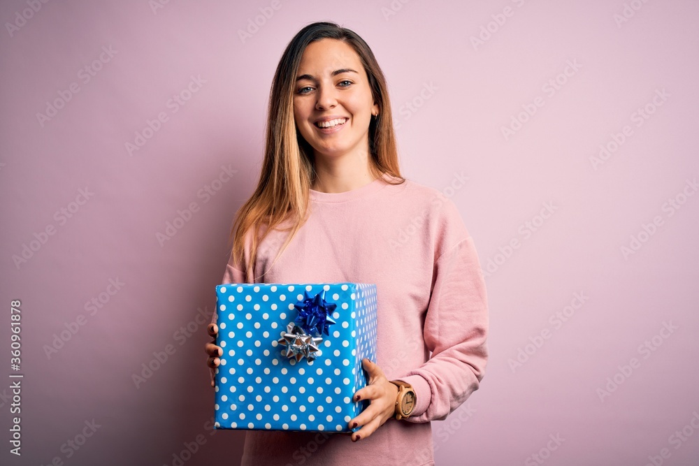 Young beautiful blonde woman with blue eyes holding birthday gift over pink background with a happy face standing and smiling with a confident smile showing teeth Young beautiful blonde woman with blue eyes holding birthday gift over pink background with a happy face standing and smiling with a confident smile showing teeth