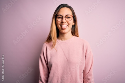 Beautiful blonde woman with blue eyes wearing sweater and glasses over pink background sticking tongue out happy with funny expression. Emotion concept.