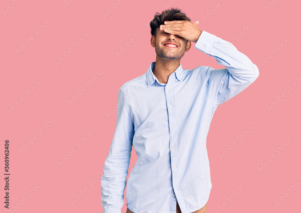 Young hispanic man wearing casual shirt smiling and laughing with hand on face covering eyes for surprise. blind concept. Young hispanic man wearing casual shirt smiling and laughing with hand on face covering eyes for surprise. blind concept.