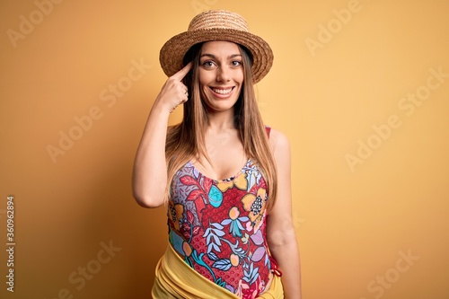 Young beautiful blonde woman wearing swimsuit and summer hat over yellow background Smiling pointing to head with one finger, great idea or thought, good memory