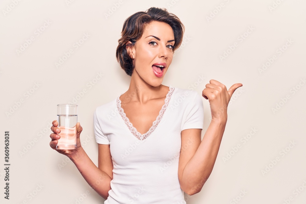 Young beautiful woman drinking glass of water pointing thumb up to the side smiling happy with open mouth Young beautiful woman drinking glass of water pointing thumb up to the side smiling happy with open mouth