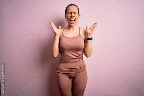 Young blonde fitness woman wearing sport workout clothes over pink isolated background celebrating mad and crazy for success with arms raised and closed eyes screaming excited. Winner concept