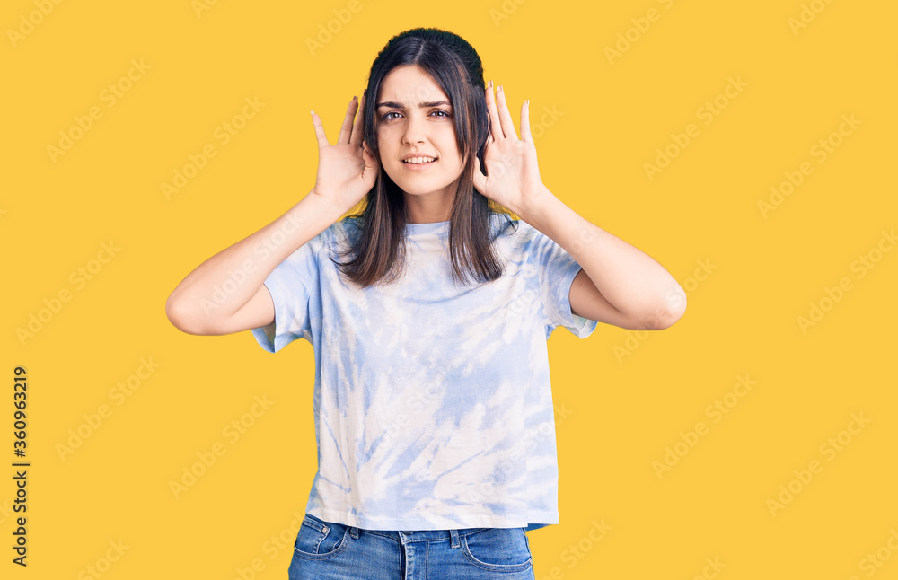 Young beautiful girl wearing casual t shirt trying to hear both hands on ear gesture, curious for gossip. hearing problem, deaf Young beautiful girl wearing casual t shirt trying to hear both hands on ear gesture, curious for gossip. hearing problem, deaf