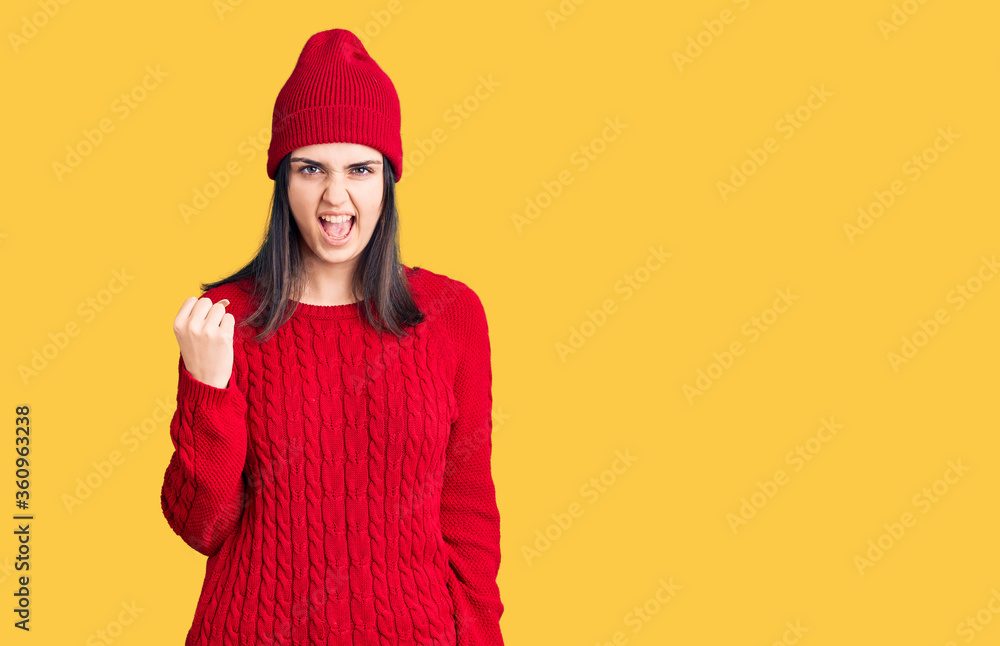 Young beautiful girl wearing sweater and wool cap angry and mad raising fist frustrated and furious while shouting with anger. rage and aggressive concept. Young beautiful girl wearing sweater and wool cap angry and mad raising fist frustrated and furious while shouting with anger. rage and aggressive concept.