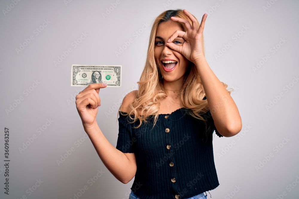 Young beautiful blonde woman holding 1 dollar banknotes over isolated white background with happy face smiling doing ok sign with hand on eye looking through fingers Young beautiful blonde woman holding 1 dollar banknotes over isolated white background with happy face smiling doing ok sign with hand on eye looking through fingers