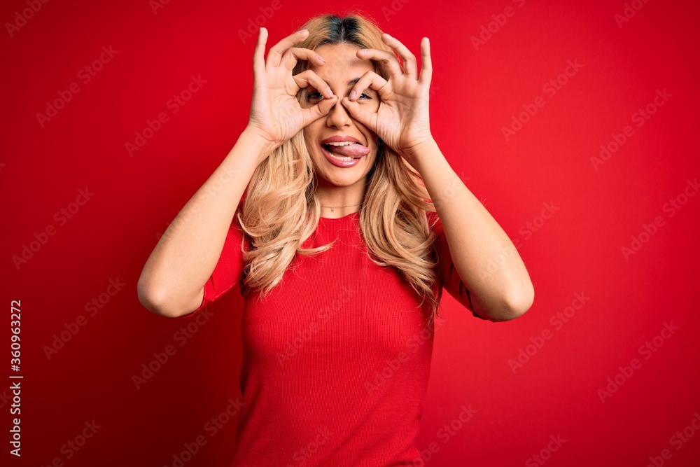 Young beautiful blonde woman wearing casual t-shirt standing over isolated red background doing ok gesture like binoculars sticking tongue out, eyes looking through fingers. Crazy expression. Young beautiful blonde woman wearing casual t-shirt standing over isolated red background doing ok gesture like binoculars sticking tongue out, eyes looking through fingers. Crazy expression.
