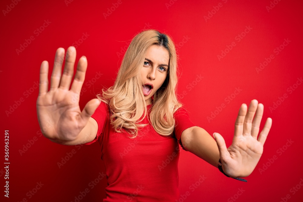 Young beautiful blonde woman wearing casual t-shirt standing over isolated red background doing stop gesture with hands palms, angry and frustration expression