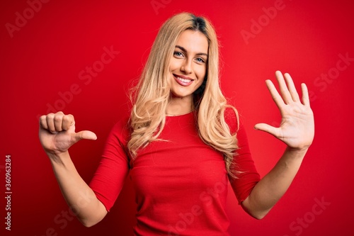 Young beautiful blonde woman wearing casual t-shirt standing over isolated red background showing and pointing up with fingers number six while smiling confident and happy.