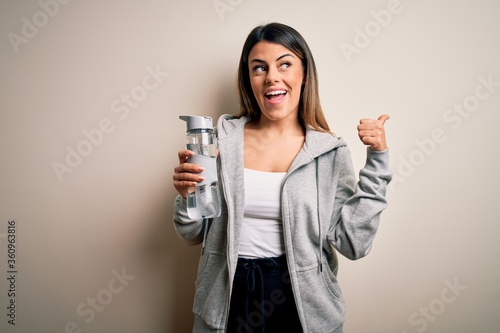 Young beautiful brunette sporty woman drinking bottle of water over isolated white background pointing and showing with thumb up to the side with happy face smiling