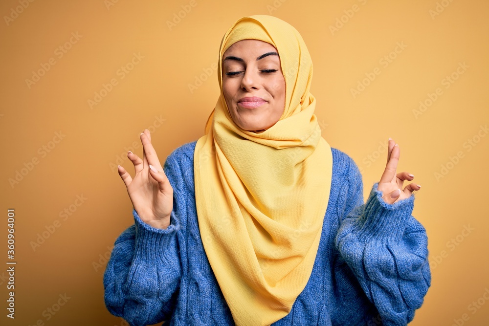 Young beautiful brunette muslim woman wearing arab hijab over isolated yellow background gesturing finger crossed smiling with hope and eyes closed. Luck and superstitious concept. Young beautiful brunette muslim woman wearing arab hijab over isolated yellow background gesturing finger crossed smiling with hope and eyes closed. Luck and superstitious concept.