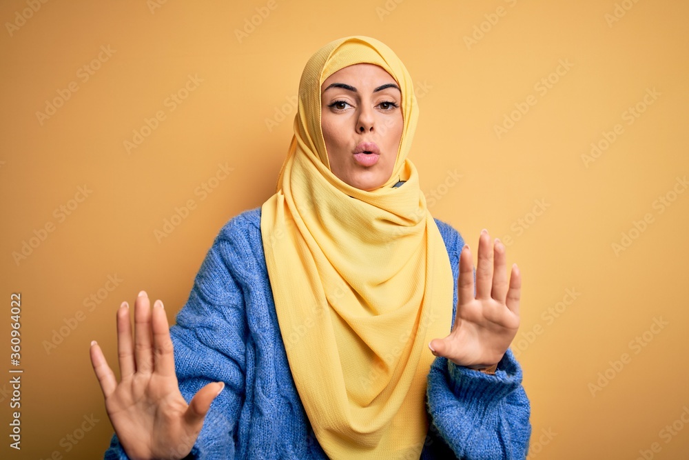Young beautiful brunette muslim woman wearing arab hijab over isolated yellow background Moving away hands palms showing refusal and denial with afraid and disgusting expression. Stop and forbidden. Young beautiful brunette muslim woman wearing arab hijab over isolated yellow background Moving away hands palms showing refusal and denial with afraid and disgusting expression. Stop and forbidden.