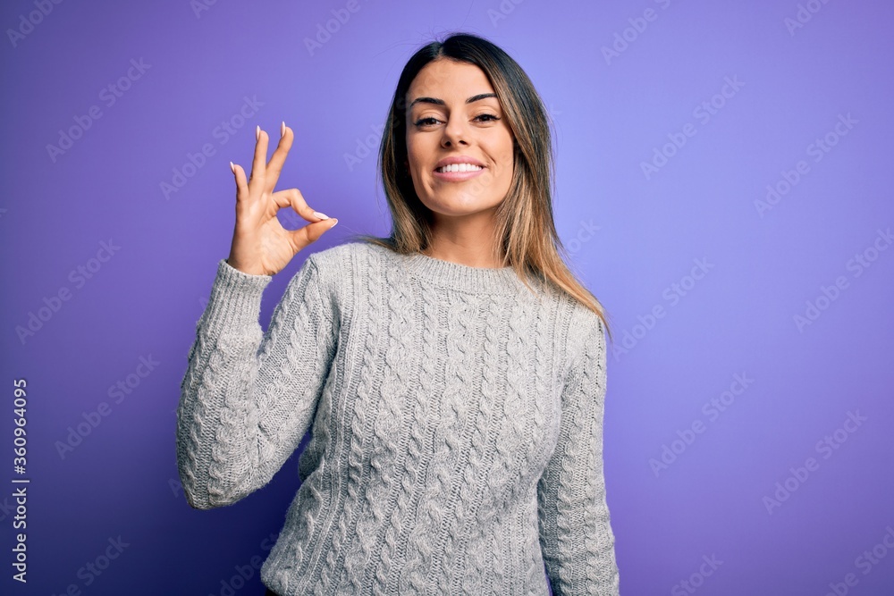 Young beautiful woman wearing casual sweater standing over isolated purple background smiling positive doing ok sign with hand and fingers. Successful expression. Young beautiful woman wearing casual sweater standing over isolated purple background smiling positive doing ok sign with hand and fingers. Successful expression.
