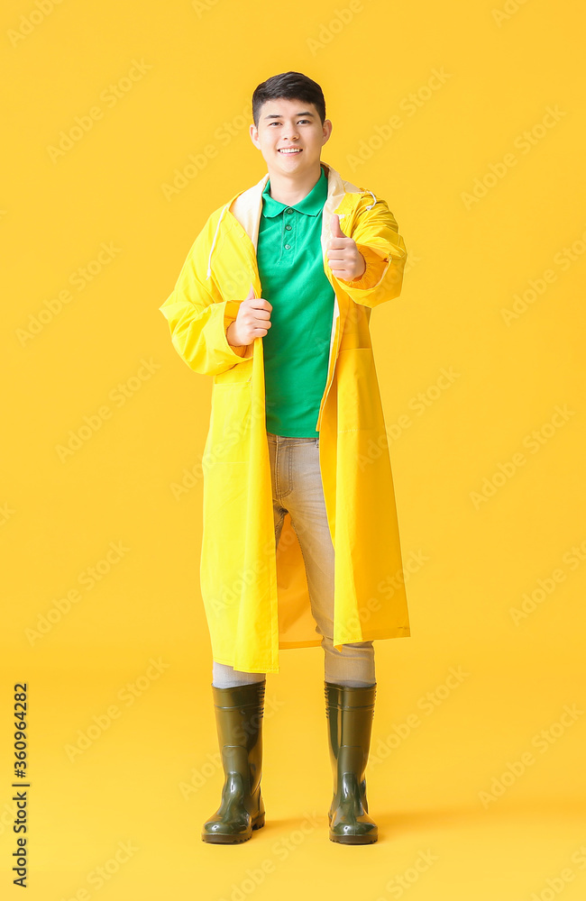 Young Asian man in raincoat showing thumb-up on color background Young Asian man in raincoat showing thumb-up on color background