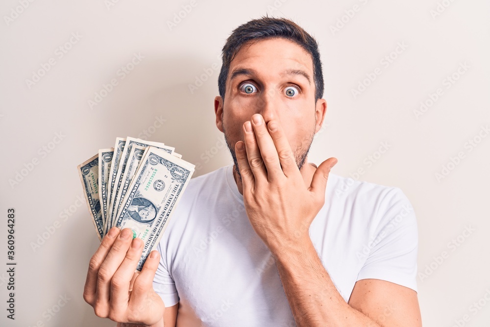 Young handsome man holding bunch of dollars banknotes over isolated white background covering mouth with hand, shocked and afraid for mistake. Surprised expression
