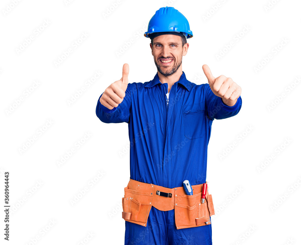 Young handsome man wearing worker uniform and hardhat approving doing positive gesture with hand, thumbs up smiling and happy for success. winner gesture. Young handsome man wearing worker uniform and hardhat approving doing positive gesture with hand, thumbs up smiling and happy for success. winner gesture.