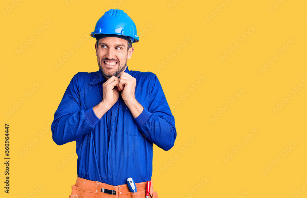 Young handsome man wearing worker uniform and hardhat laughing nervous and excited with hands on chin looking to the side